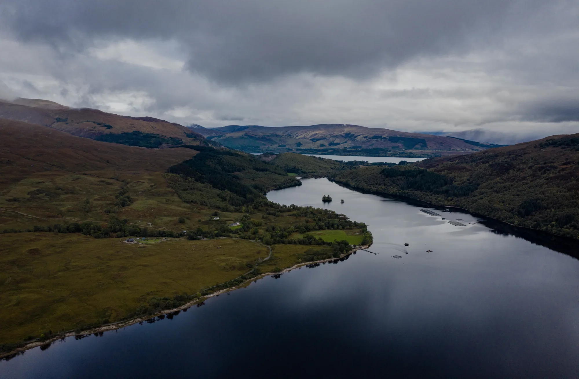 An aerial view along a still Scottish loch between low hills under heavy cloud